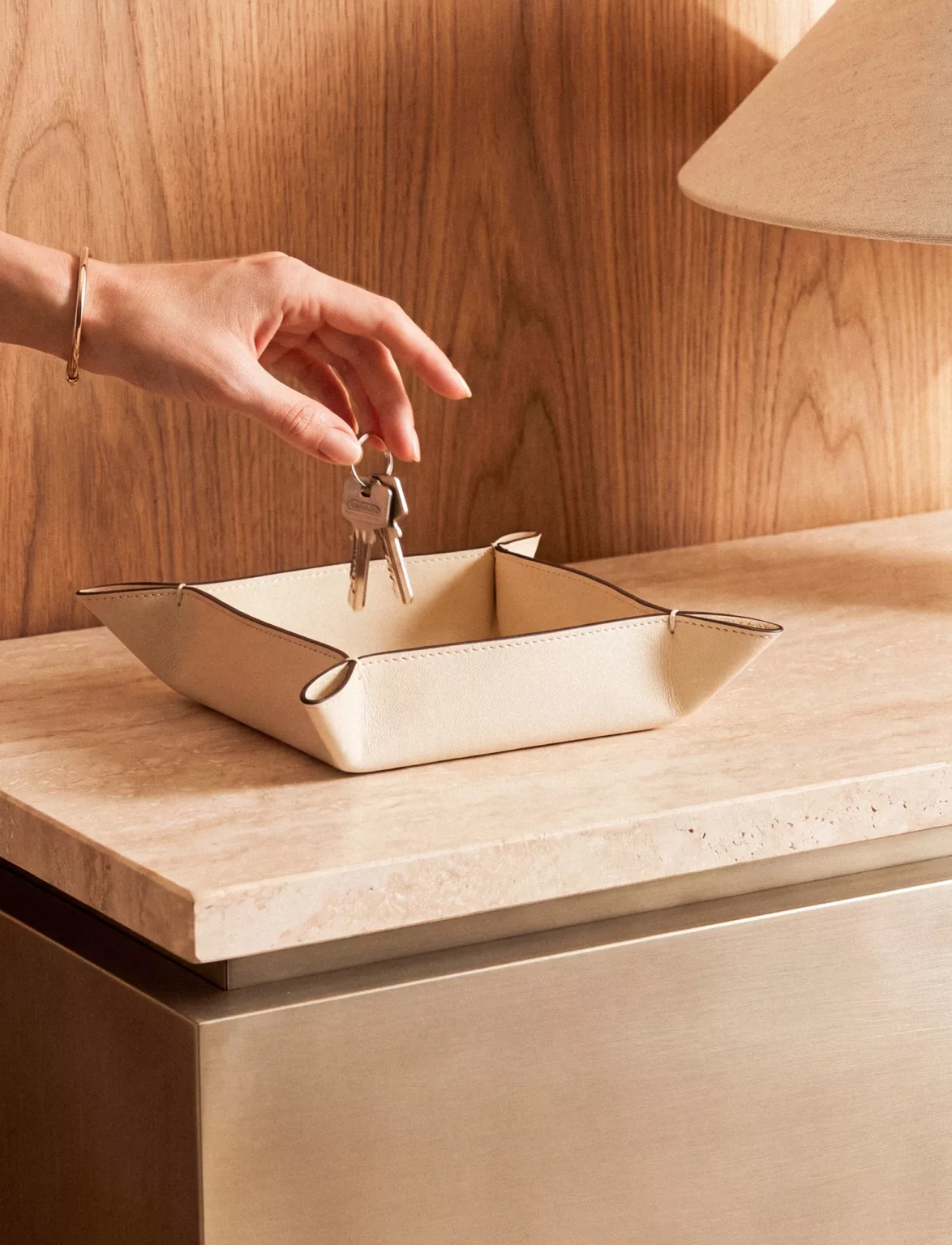 Une main dépose un trousseau de clés dans un vide-poche en cuir crème, sur un meuble en pierre claire.