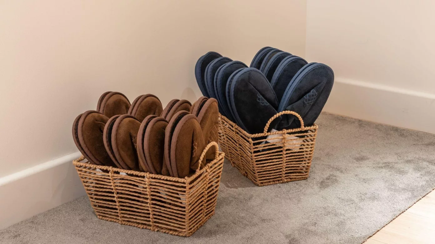Two woven baskets filled with brown and navy felt slippers, in the corner of a bedroom.