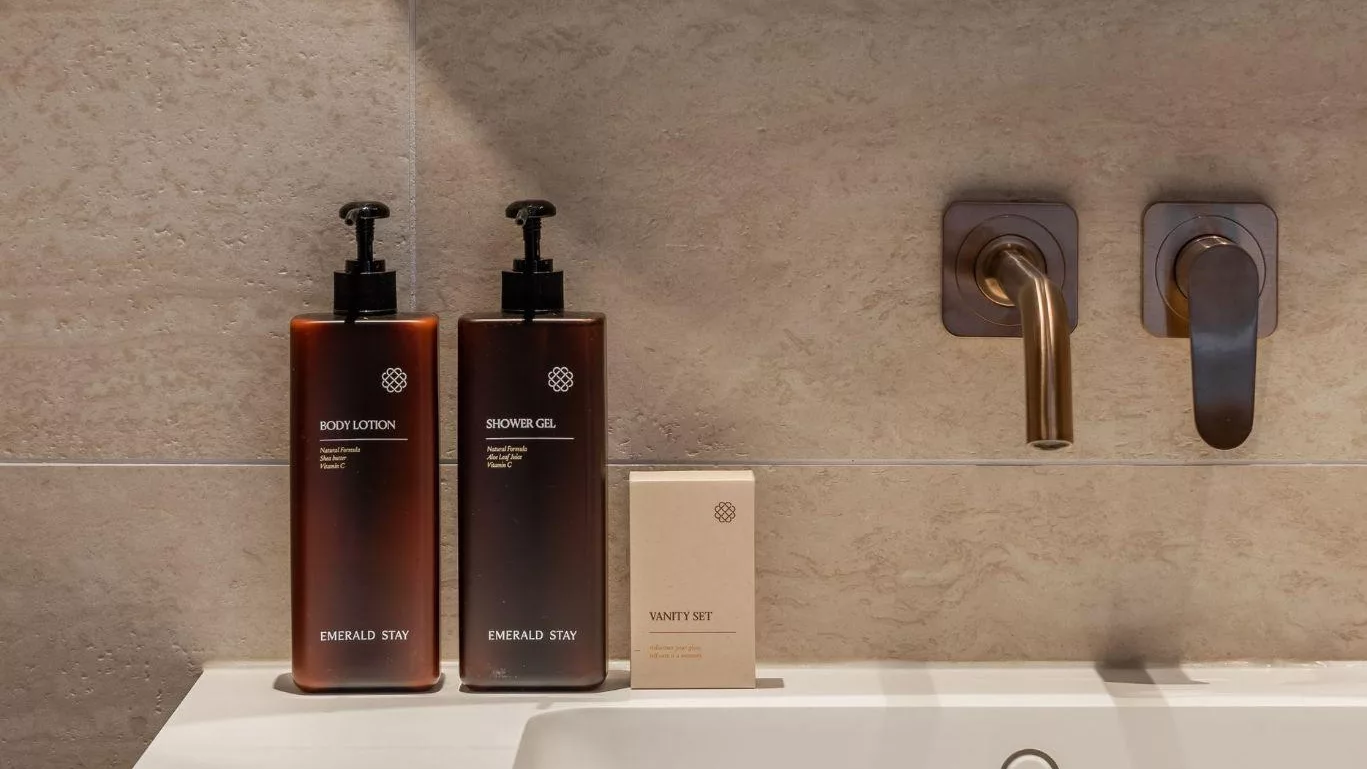 Two amber bottles and an Emerald Stay vanity set on a basin, against a pale stone wall.