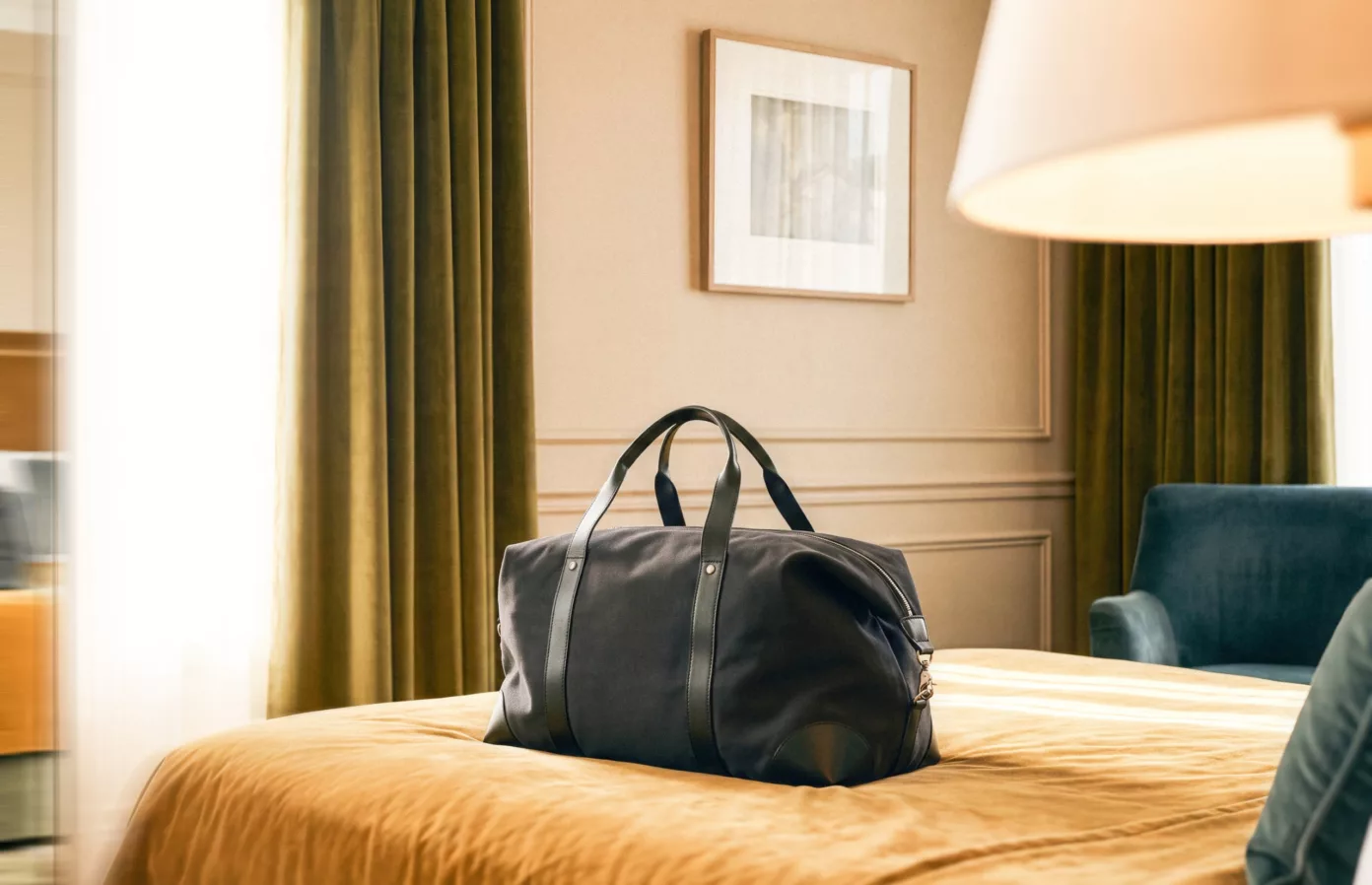A black travel bag with leather handles on the bed of a hotel room, in the glow of a lamp.