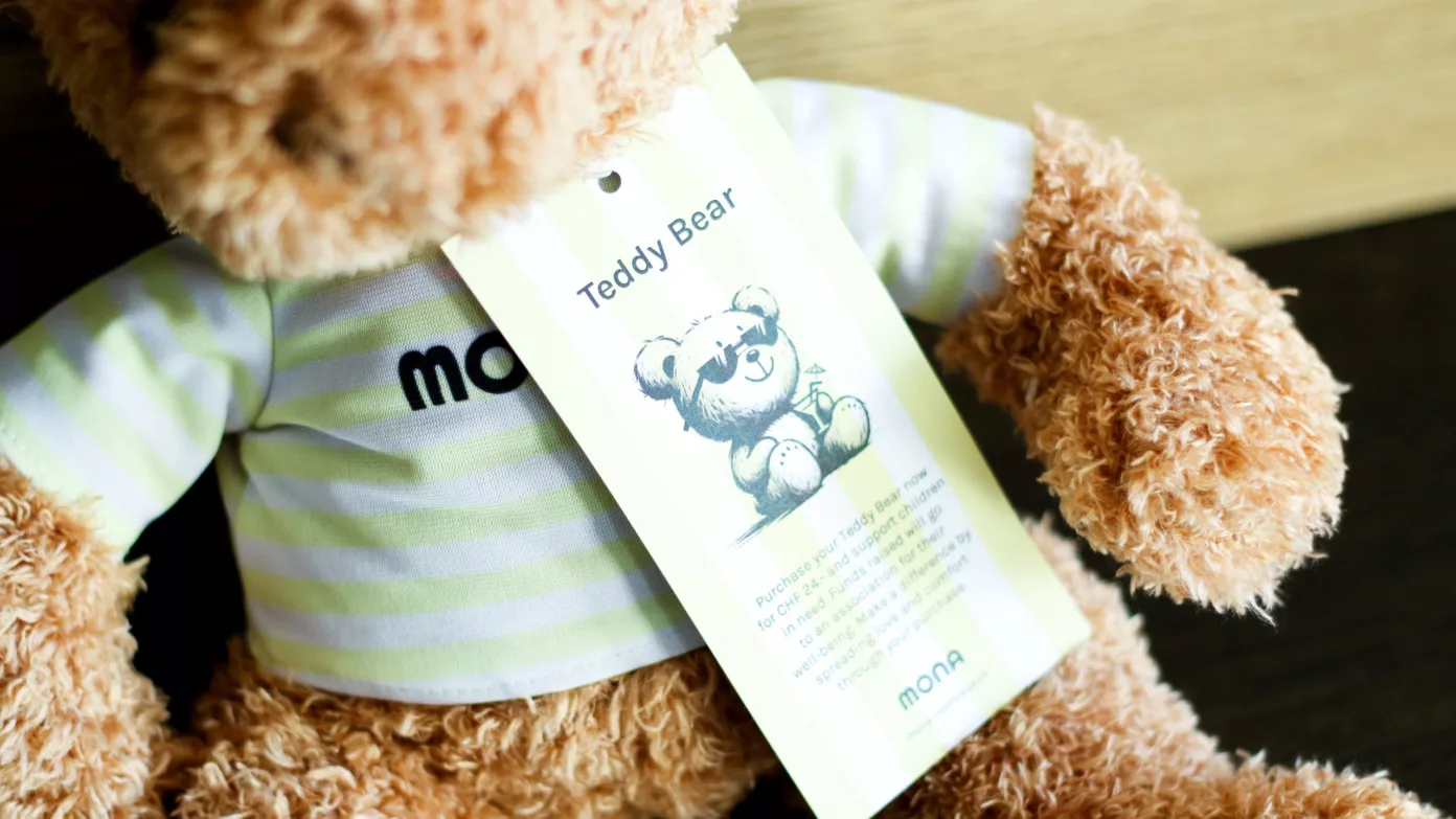 A teddy bear wearing a striped MONA T-shirt, with its Teddy Bear tag.