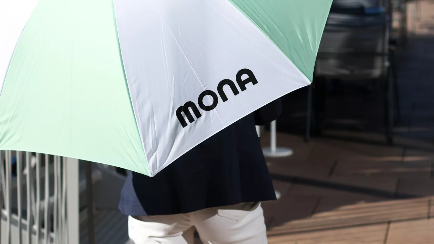 A guest under a mint and white MONA umbrella, on a jetty.