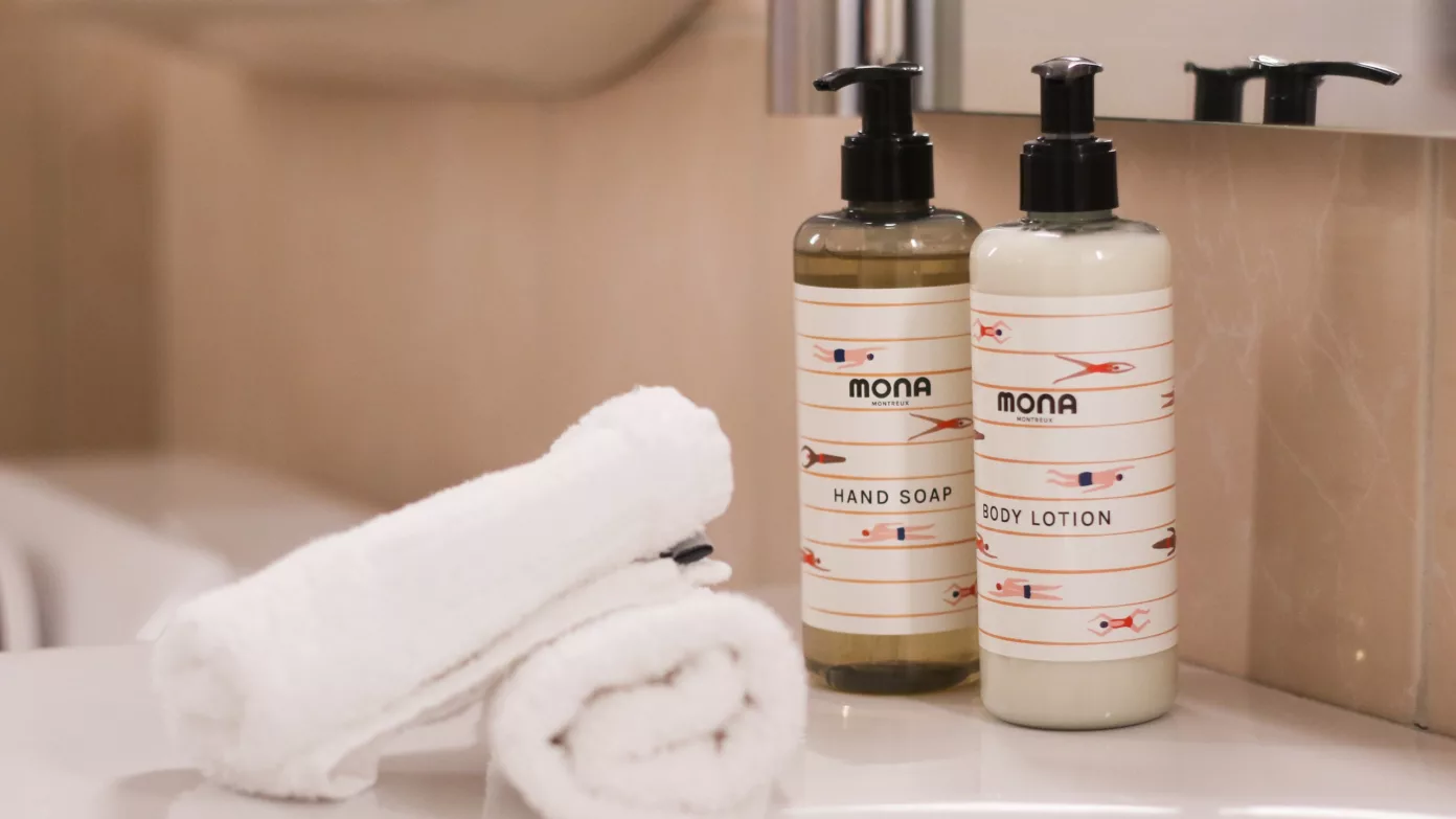 MONA hand soap and body lotion beside a rolled towel, on the edge of a bathtub.