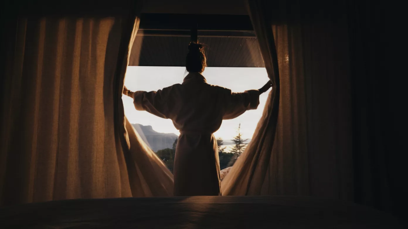 Une voyageuse en peignoir ouvre les rideaux de sa chambre sur les montagnes.