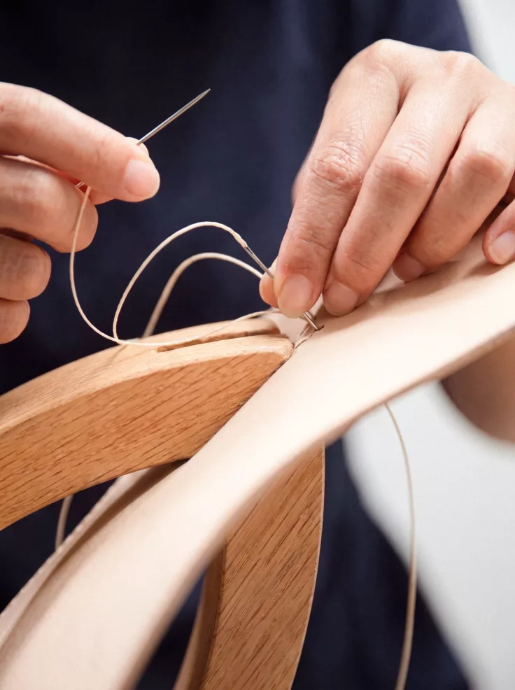 Des mains cousent au fil ciré une bande de cuir naturel sur une pièce de bois.