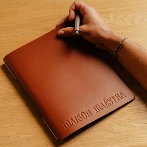 Une main tient un stylo sur un carnet en cuir cognac marqué Maison Maëstra, posé sur une table en chêne.
