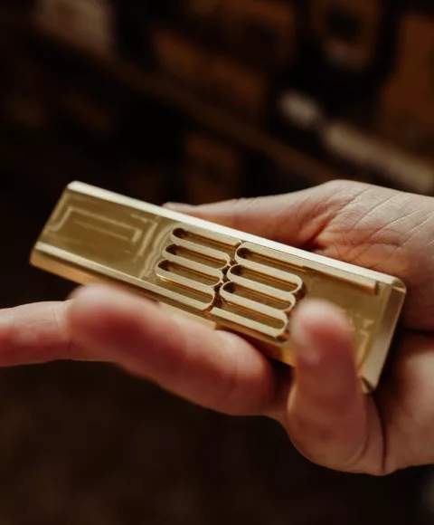 A hand holding a brass stamping die engraved with a raised pattern, in the half-light of the workshop.