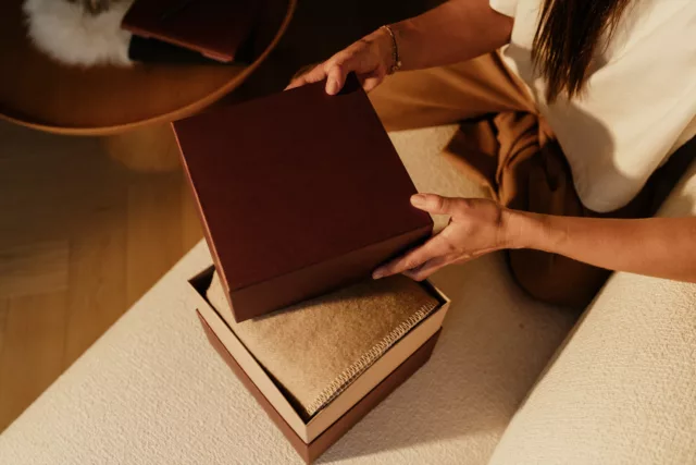 Deux mains soulèvent le couvercle d’un coffret bordeaux garni d’un plaid beige.