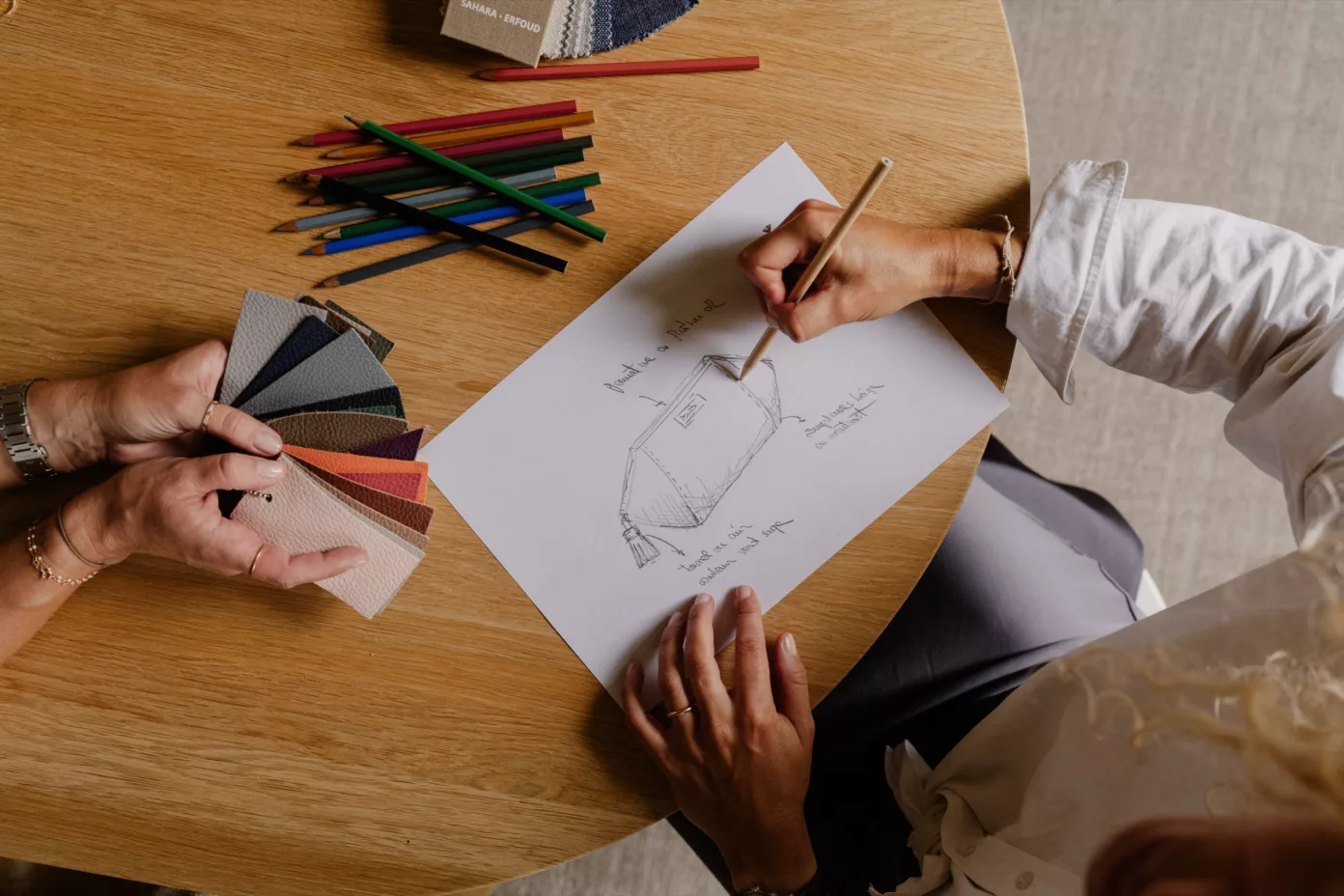 Deux mains esquissent un objet au crayon, près d’un nuancier de cuirs et de crayons de couleur.