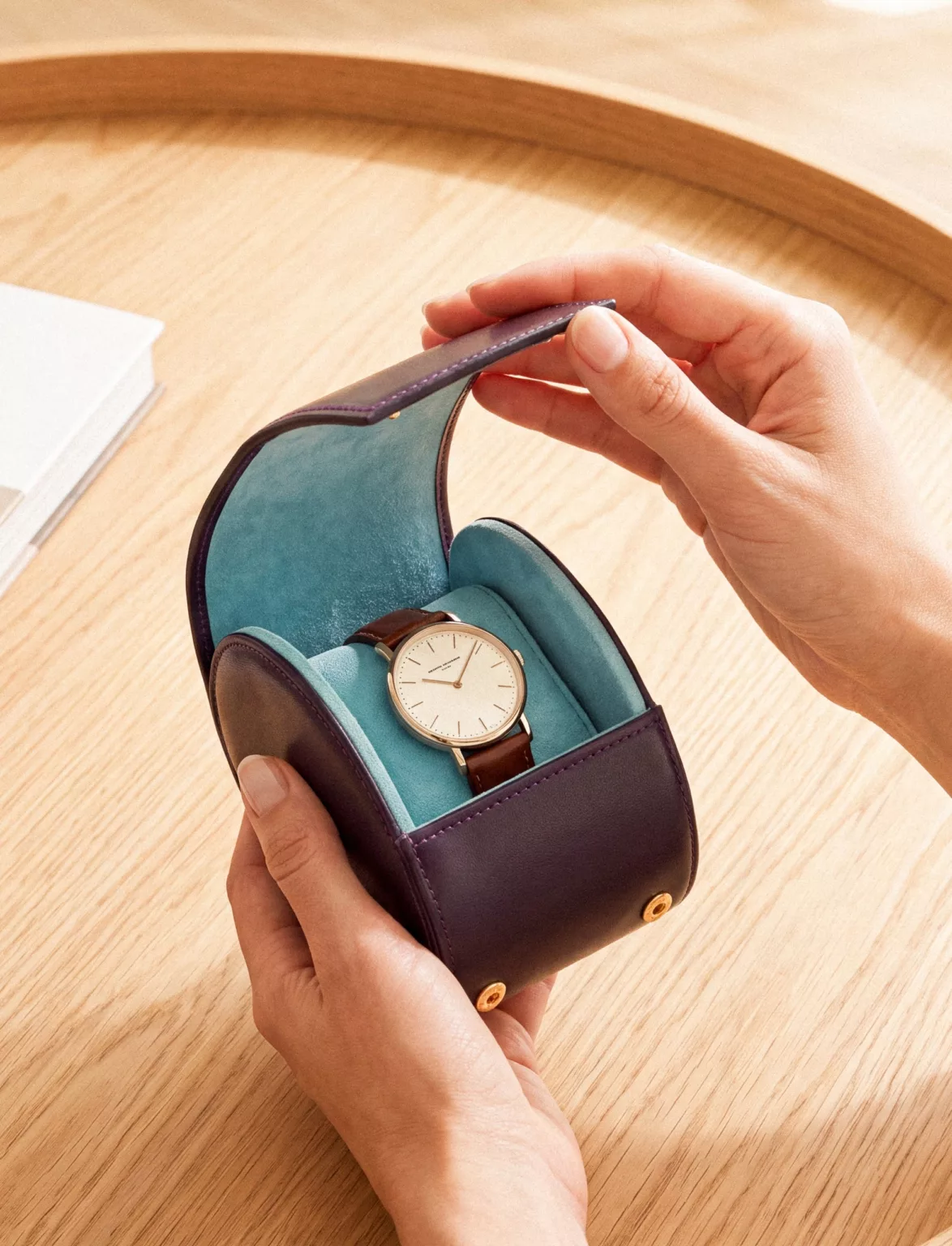 Des mains ouvrent un écrin de voyage prune doublé de velours turquoise, qui abrite une montre.