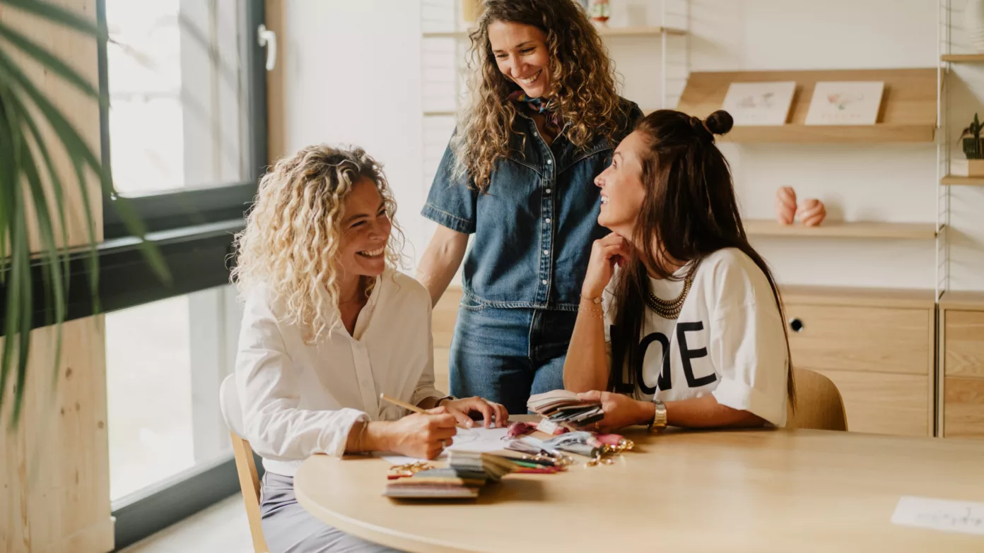 Three women share a smile around a studio table covered with colour swatches and samples.