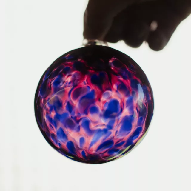 Blown glass bauble with blue, pink and violet swirls, held up against the light, bespoke lifestyle piece