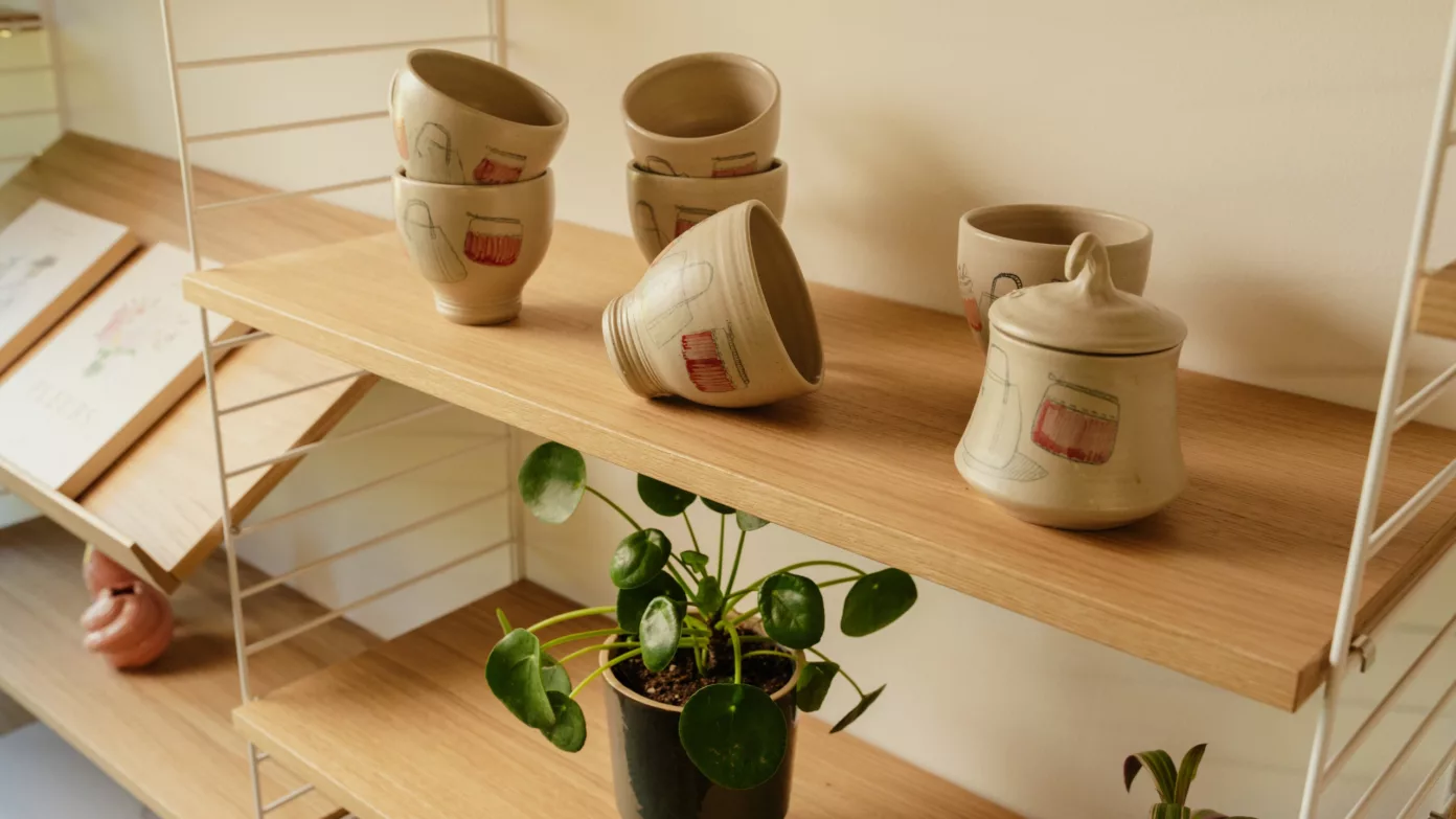 Des tasses et un sucrier en céramique peinte à la main sur une étagère en bois clair, au-dessus d’une plante.