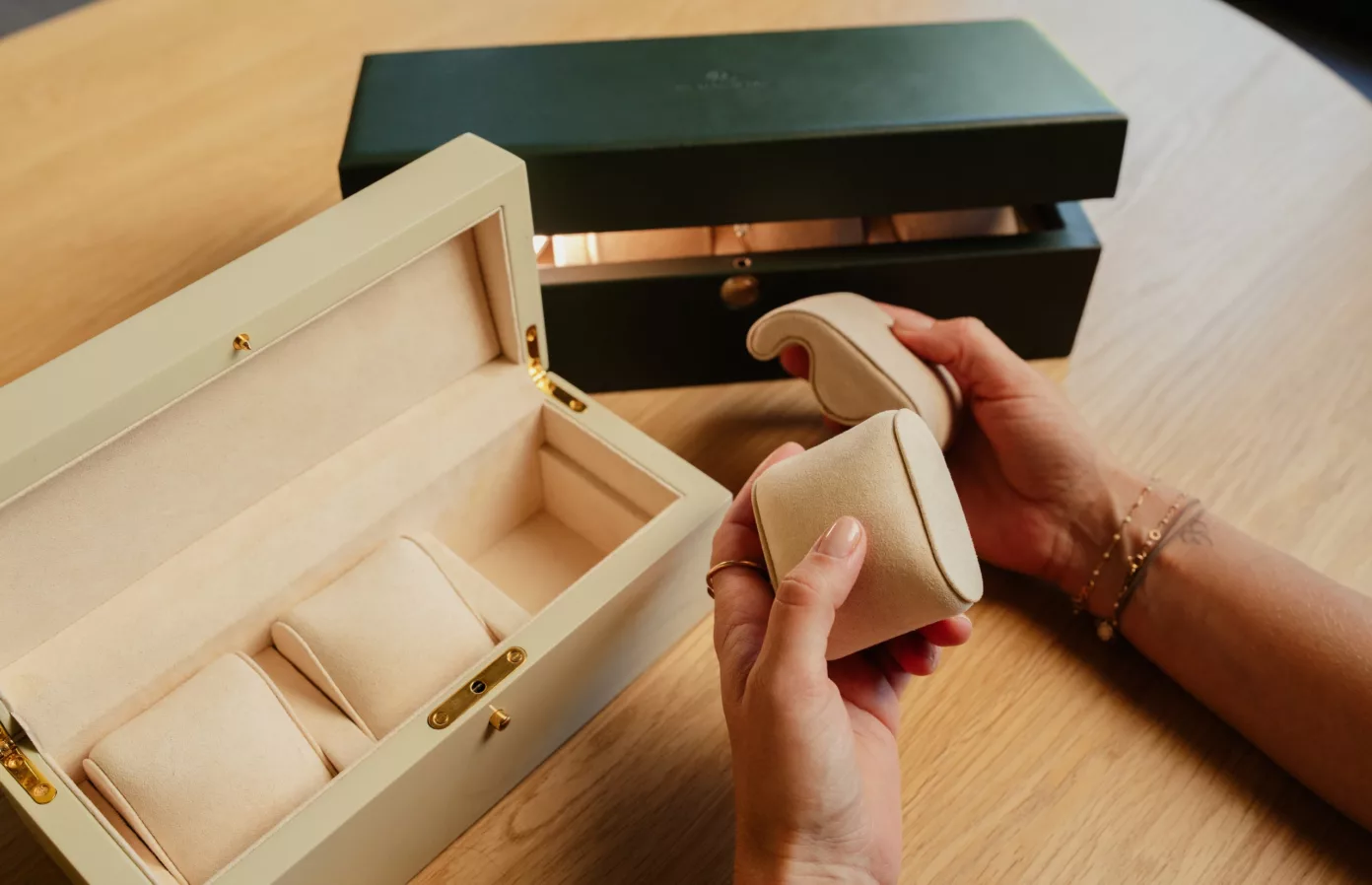 Mains tenant deux coussins de montre devant des coffrets crème et vert ouverts, écrins de montres sur-mesure, Maison Maëstra