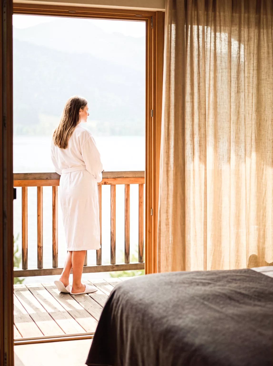 Une voyageuse en peignoir, sur le seuil du balcon, contemple un lac et les montagnes.