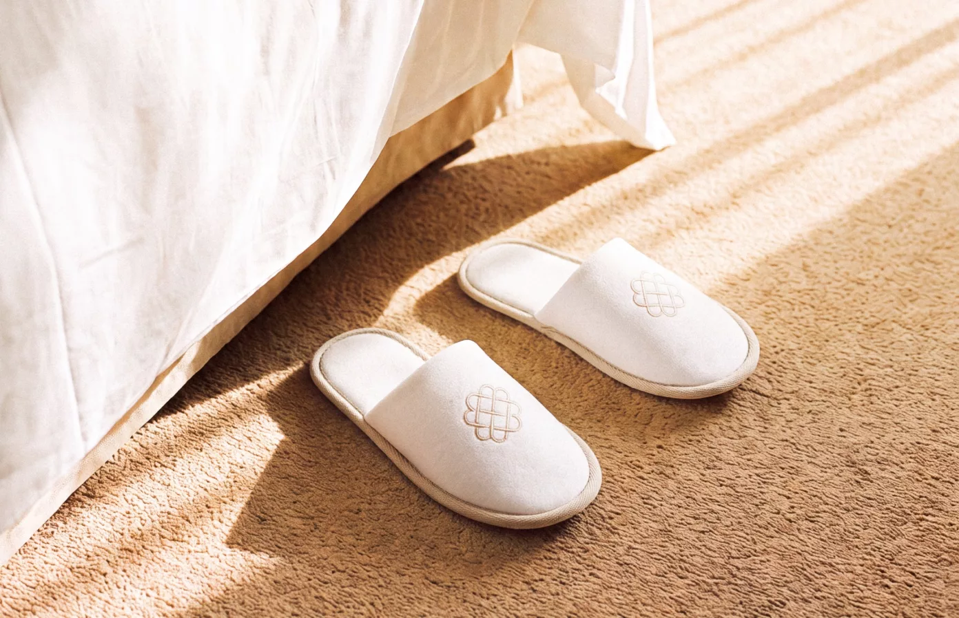Embroidered white slippers at the foot of a sunlit hotel bed, bespoke guest amenities by Maison Maëstra