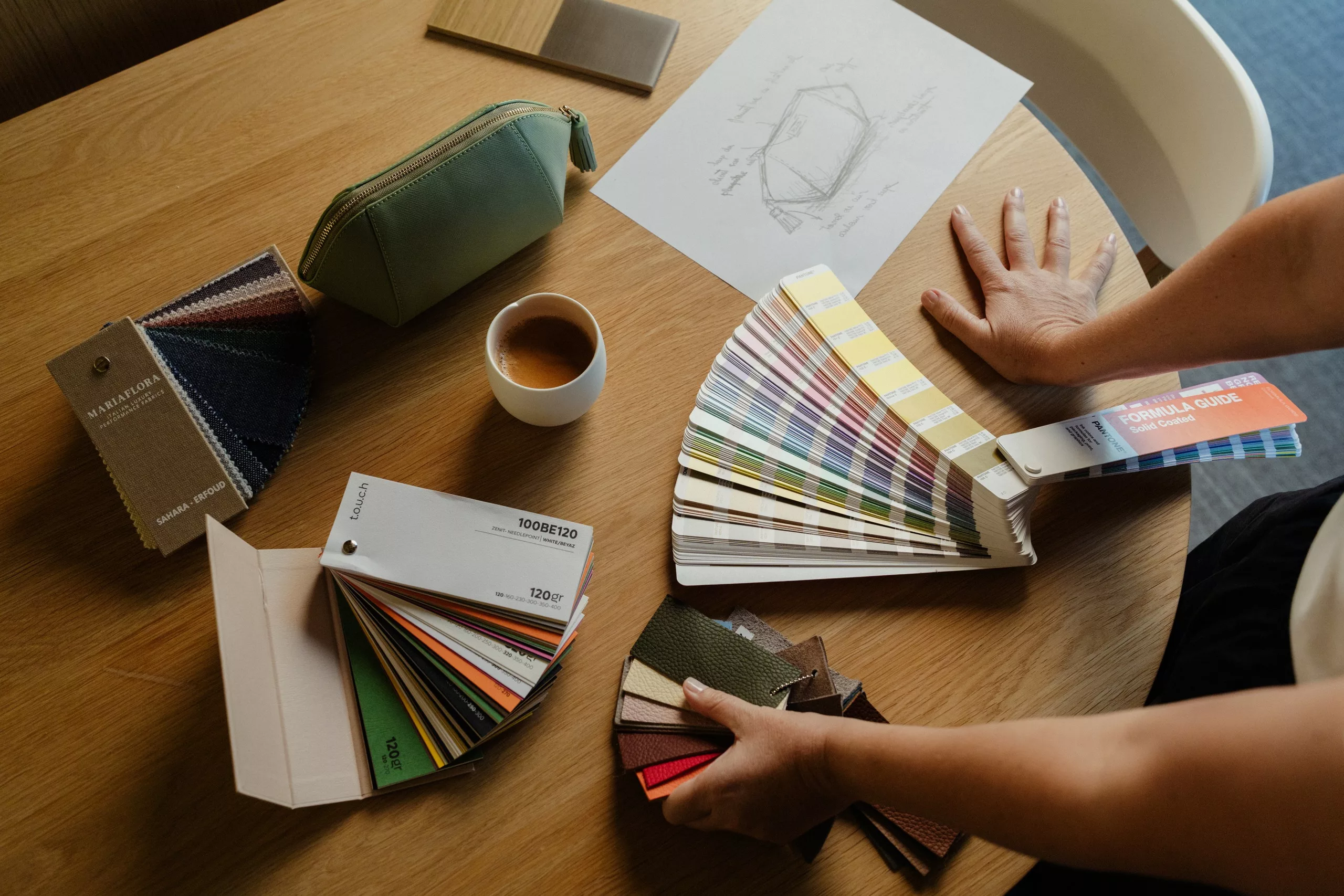 Des mains parcourent des nuanciers de couleurs et des échantillons de cuir autour d’un croquis, d’une tasse de café et d’une trousse verte, sur une table en chêne.