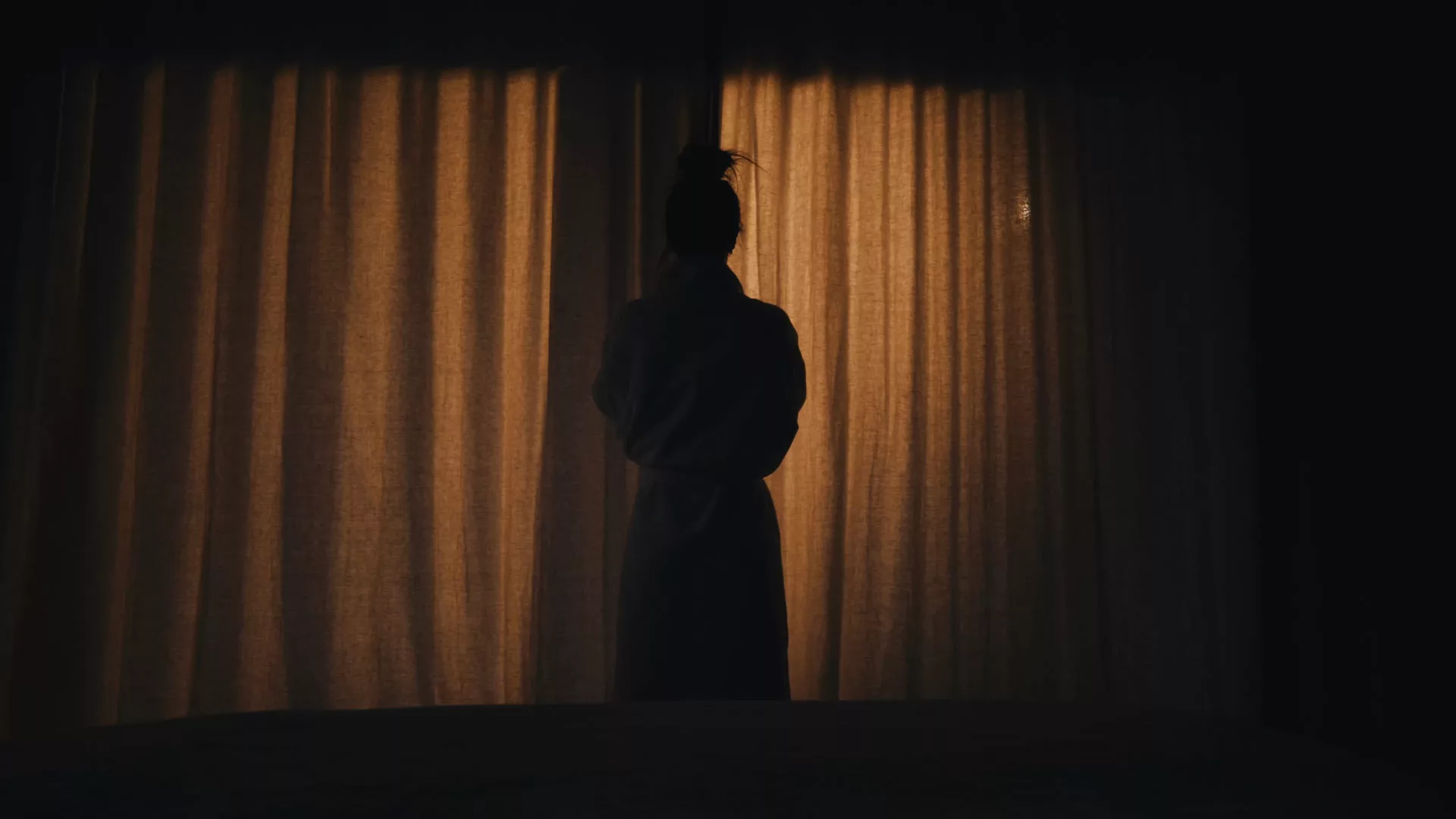 A woman’s silhouette against the closed curtains of a bedroom, backlit.